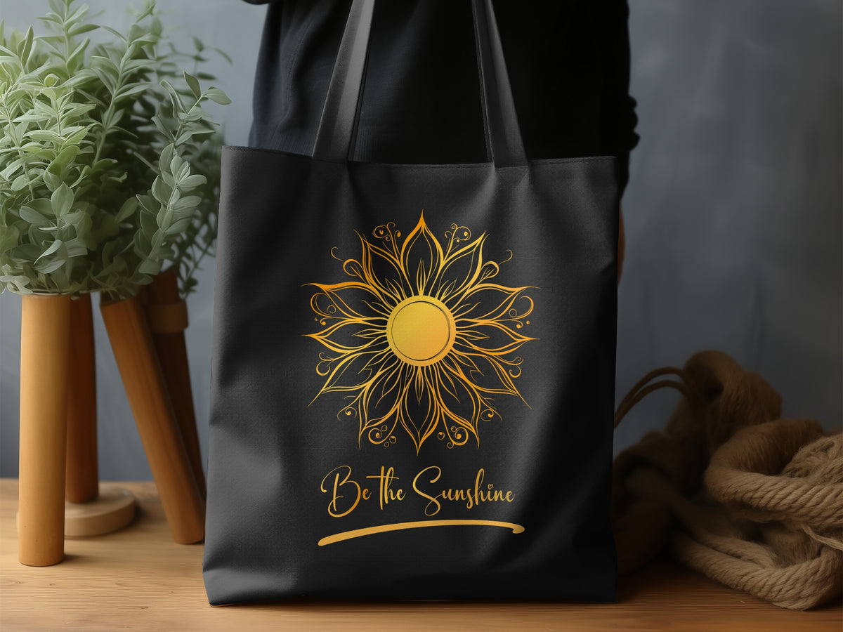 Black canvas Be The Sunshine Tote Bag lifestyle display featuring a radiant golden mandala sun, ornate petal detailing and flowing Be the Sunshine script beneath.