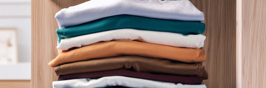 Folded men's t-shirts in blue, teal, white, orange, brown and burgundy on an oak wardrobe shelf.