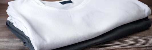 Stack of folded crew-neck t-shirts in white, charcoal grey and black on a dark timber surface.
