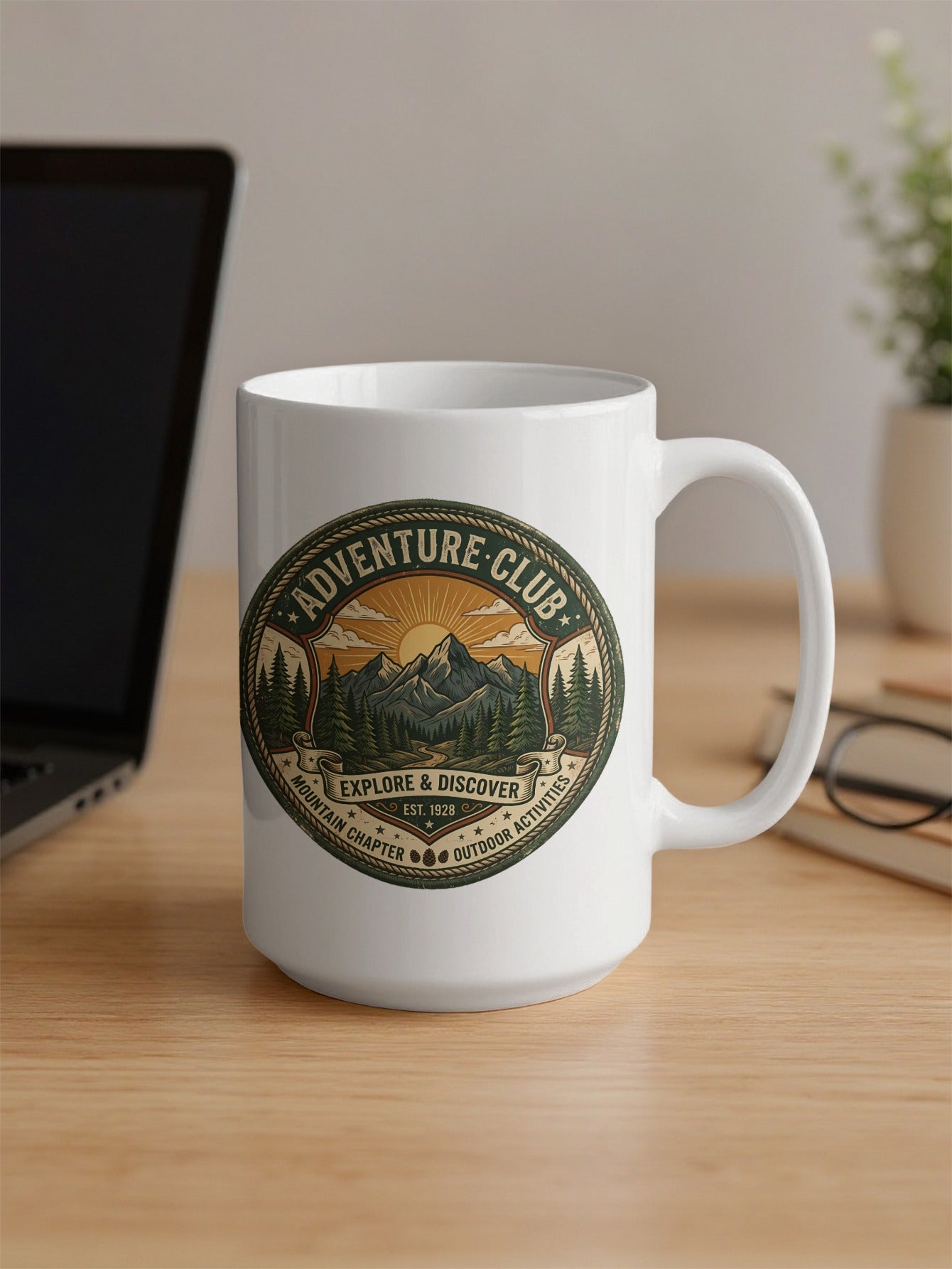 Adventure Club 15oz mug on office desk featuring twin mountains, forest trail and vintage outdoor circular badge.