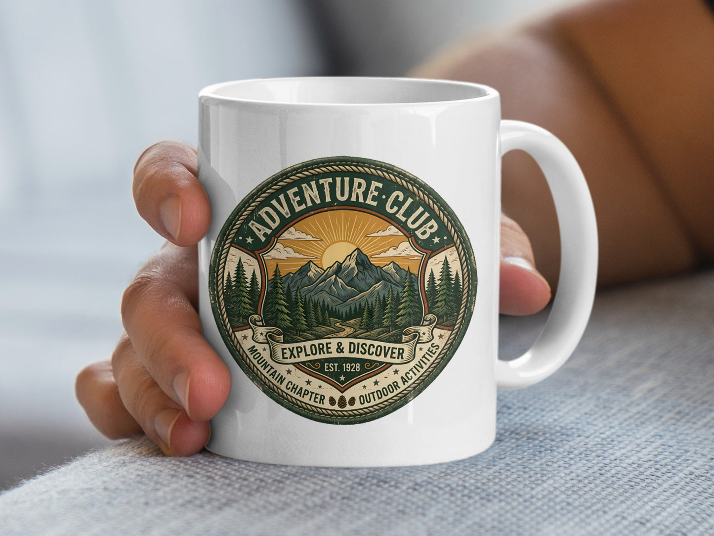 Hand holding Adventure Club ceramic mug with Explore & Discover banner and mountain landscape artwork.