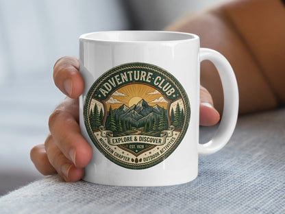 Hand holding Adventure Club ceramic mug with Explore & Discover banner and mountain landscape artwork.