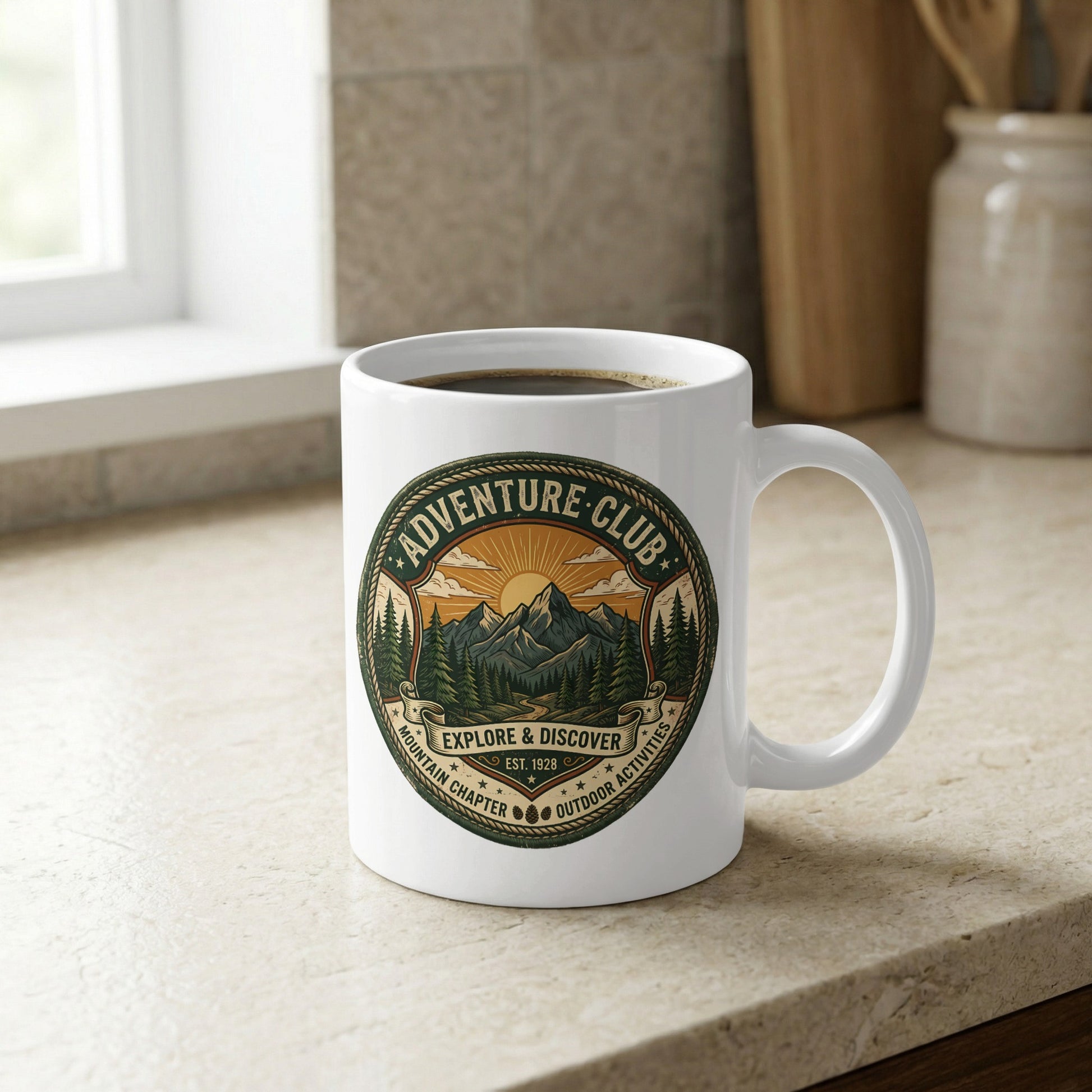 Adventure Club 11oz mug displayed on kitchen counter featuring pine forest, mountain trail and sunburst circular artwork.