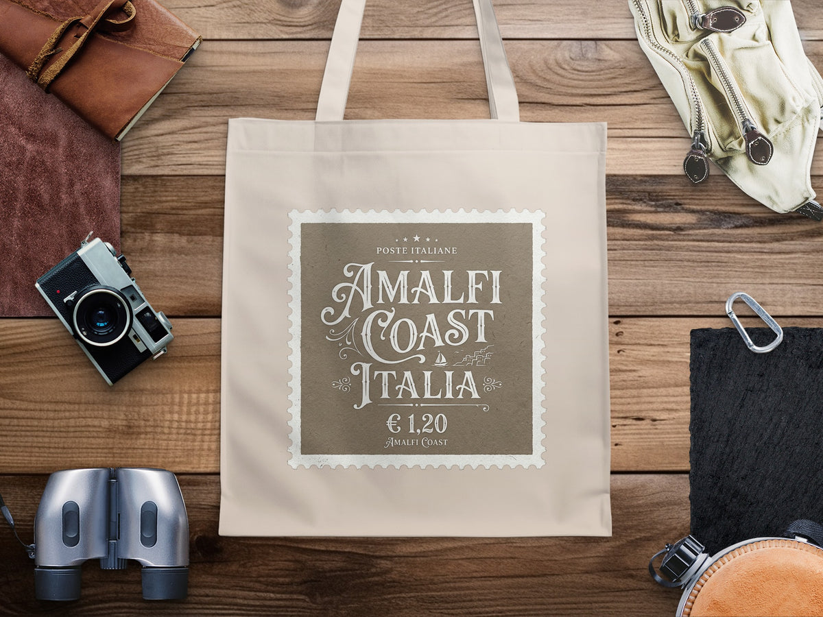 Amalfi Coast Italia Tote Bag on a wooden table, featuring ornate lettering, small sailboat icon and vintage postal-inspired details.