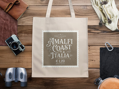 Amalfi Coast Italia Tote Bag on a wooden table, featuring ornate lettering, small sailboat icon and vintage postal-inspired details.