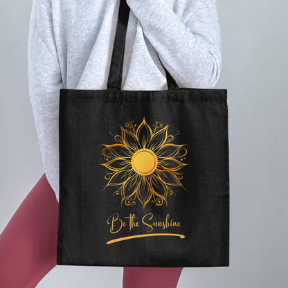 Be The Sunshine black tote bag held by a woman, showing ornate golden sun detailing, relaxed gold script lettering and soft underline accent.