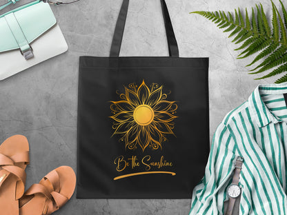 Be The Sunshine black canvas tote bag laid flat with a symmetrical golden mandala sun featuring layered petal forms radiating around its centre and gold script lettering.