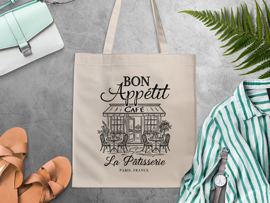 Black line-art Parisian café design with rattan-style chairs, outdoor bistro tables and leafy potted plants on a canvas cotton graphic tote bag.