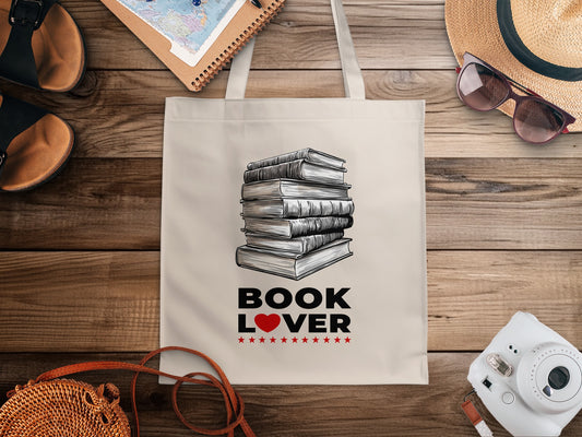 Cream Book Lover canvas tote bag with seven stacked books in detailed monochrome linework, grey shading and visible pages with black lettering and red heart accent.
