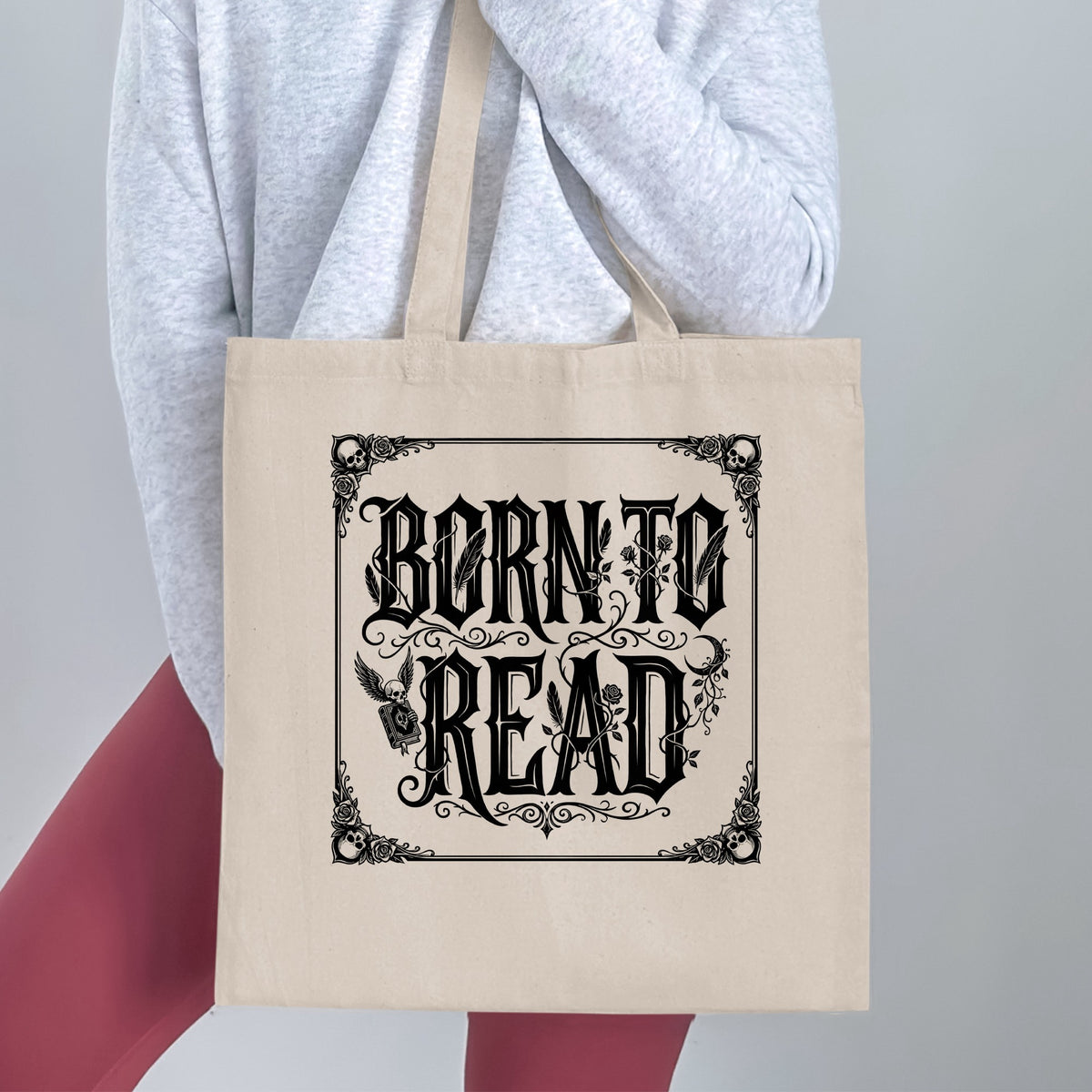 Born to Read canvas tote bag held by a woman, showing blackletter lettering, corner skulls and winged skull clutching a closed book.
