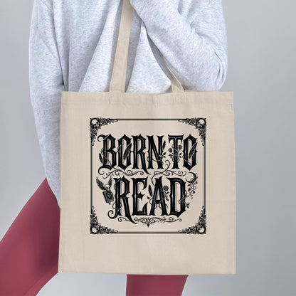 Born to Read canvas tote bag held by a woman, showing blackletter lettering, corner skulls and winged skull clutching a closed book.