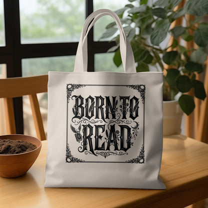 Natural-tone canvas Born to Read Tote Bag on wooden table with elaborate blackletter type, floral motifs and skulls framing the design.