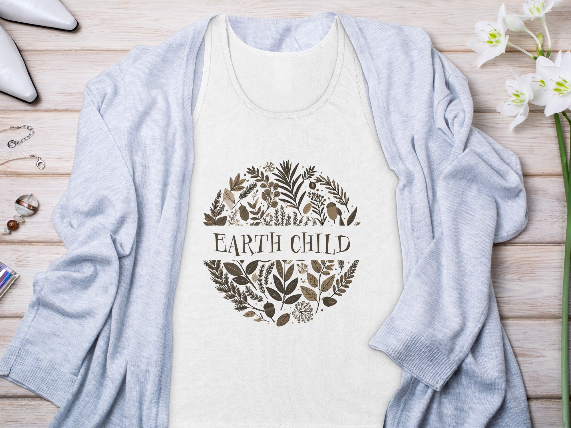White Earth Child tank top with illustrated fern fronds, seed pods, acorns, and centred serif typography styled with cardigan on wooden surface.