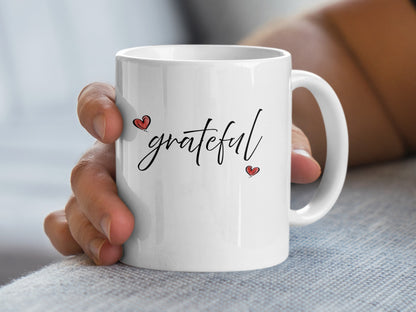 Handheld 11oz white Grateful Heart Mug showing expressive black script lettering with delicate red heart accents.