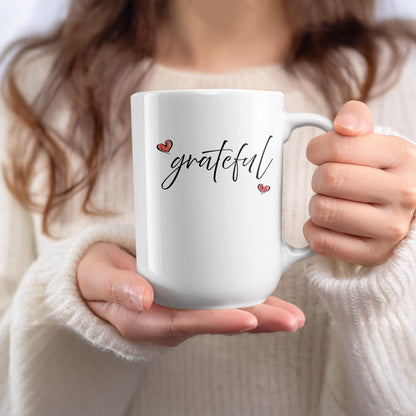 Handheld 15oz white Grateful Heart Mug featuring minimalist black script and two contrasting red heart details with black outlines.