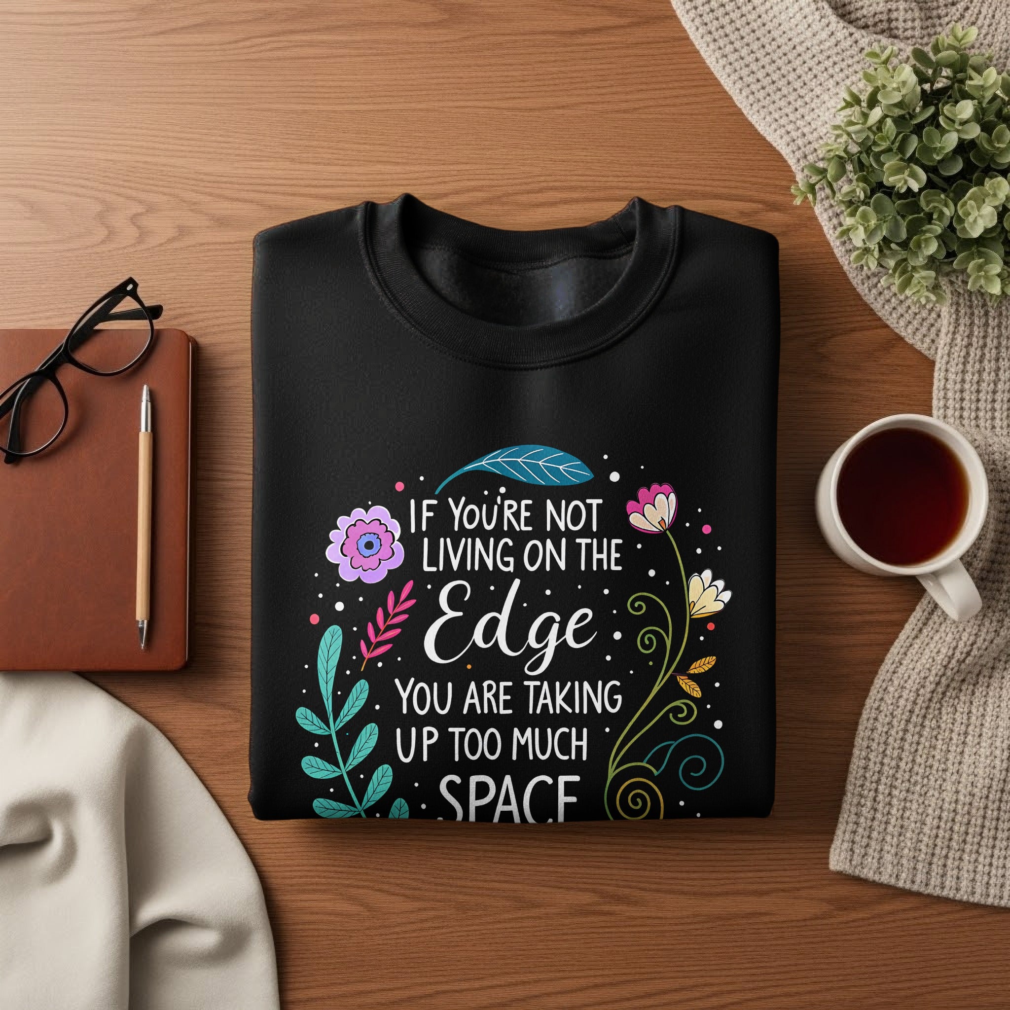 Folded Live On The Edge sweatshirt in black featuring colourful floral wreath illustration and white layered quote typography.