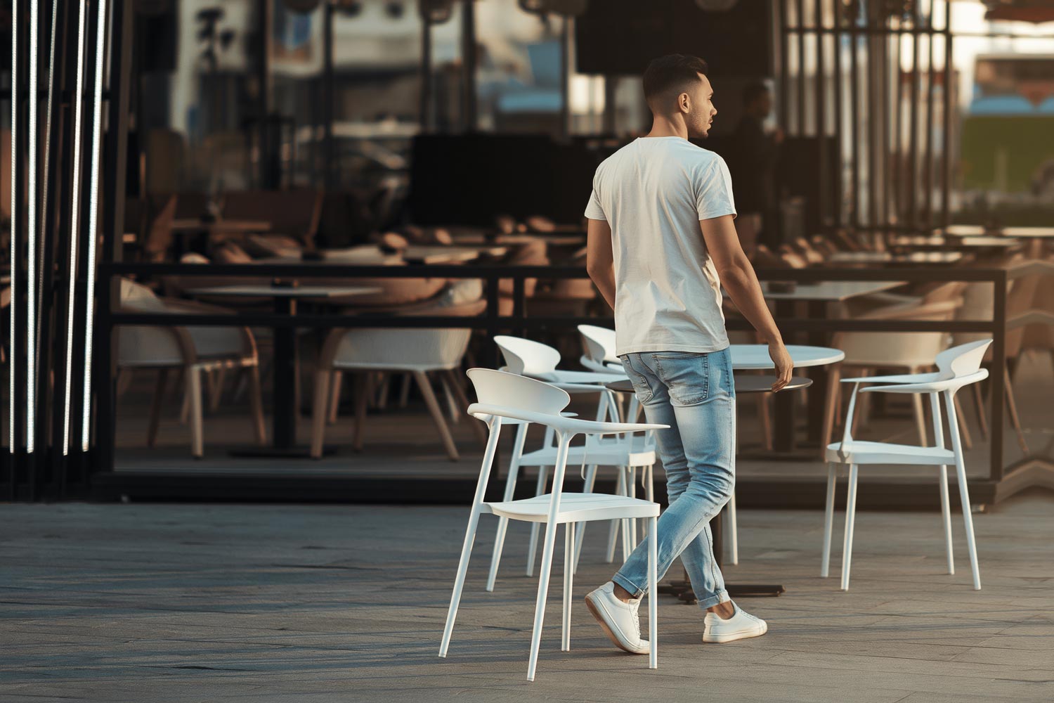 Man wearing a white crew neck tee in an outdoor cafe setting, styled with light wash jeans and white sneakers for a casual streetwear look.