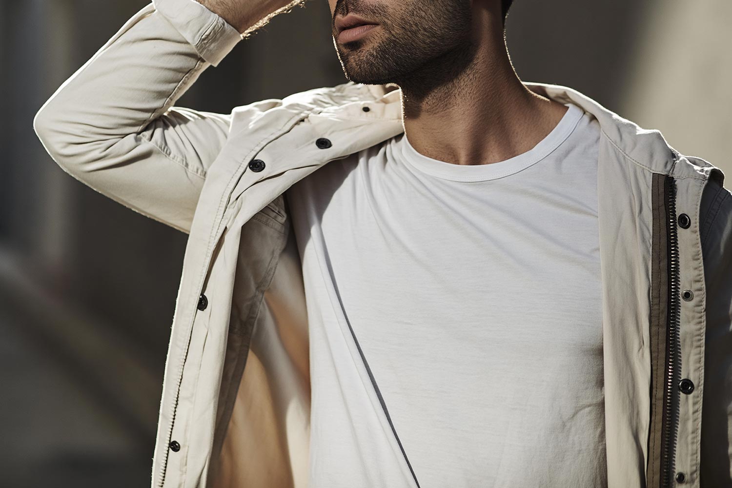 Man wearing white crew neck t-shirt layered under an open cream jacket in an outdoor setting.