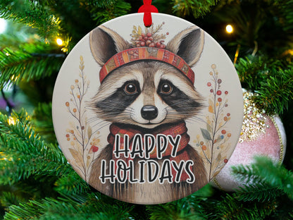 Round holiday ornament showcasing a raccoon portrait with red scarf, winter greenery, and berry accents.