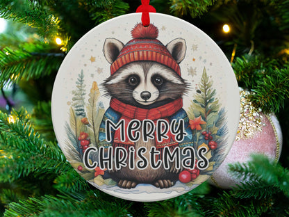 Round holiday ornament showcasing a cosy raccoon character in a snowy forest scene with seasonal greenery.