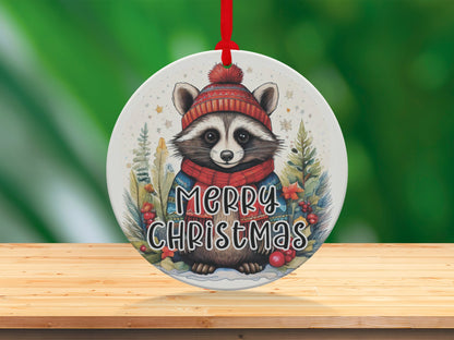 Storybook-style raccoon ornament with snow-dusted evergreens, holly details, and warm woodland holiday charm.