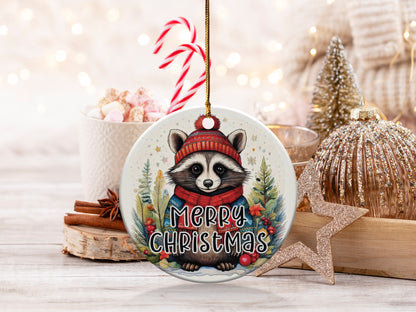 Watercolour raccoon ornament wearing a red pom-pom beanie, teal sweater, and knitted scarf among snowy pines.