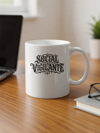 White 11oz Social Vigilante Mug on an office desk featuring bold black Victorian-style lettering and ornate framing.