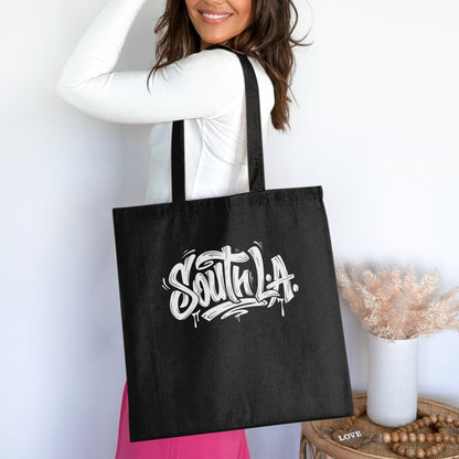 Black South L.A. Tote Bag worn over the shoulder, featuring white graffiti lettering, paint drips and dark shadowing.