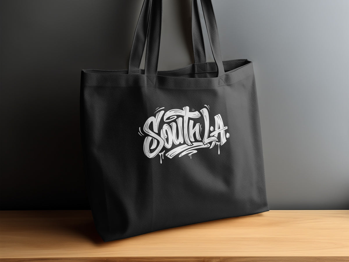 South L.A. Tote Bag displayed on a sideboard, showing white graffiti lettering with paint drips and flourishes.