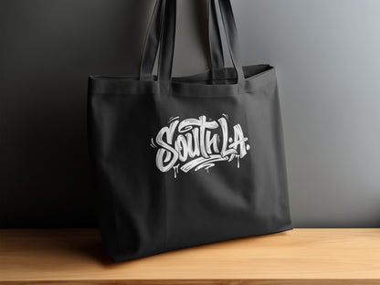 South L.A. Tote Bag displayed on a sideboard, showing white graffiti lettering with paint drips and flourishes.
