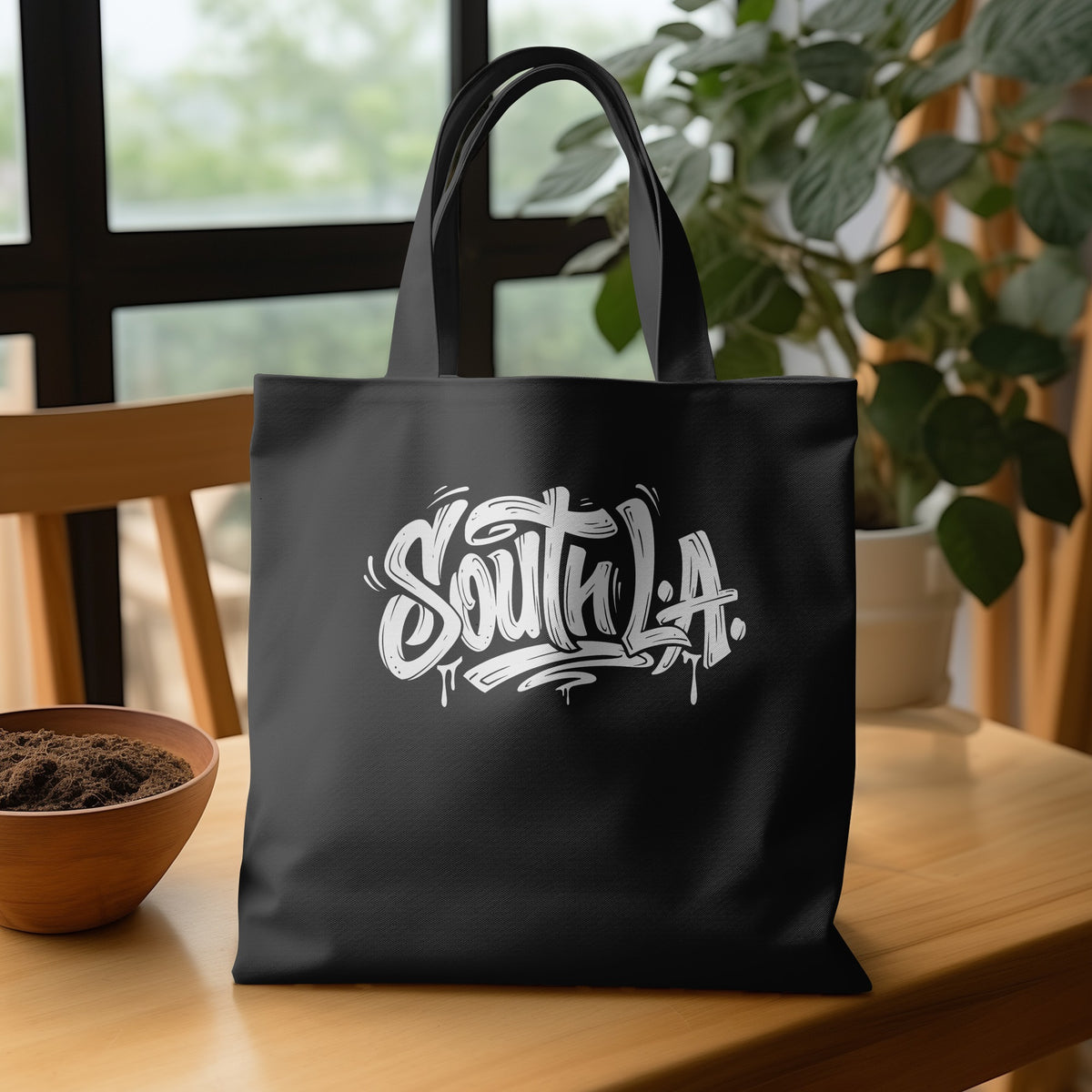 Black canvas South L.A. Tote Bag on a wooden table, showing white lettering, dark shadowing and dripping paint details.
