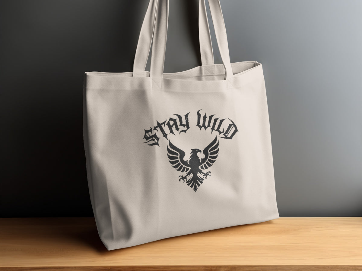 Stay Wild Heraldic Eagle Tote Bag displayed on a sideboard, showing the arched blackletter lettering and symmetrical eagle emblem.